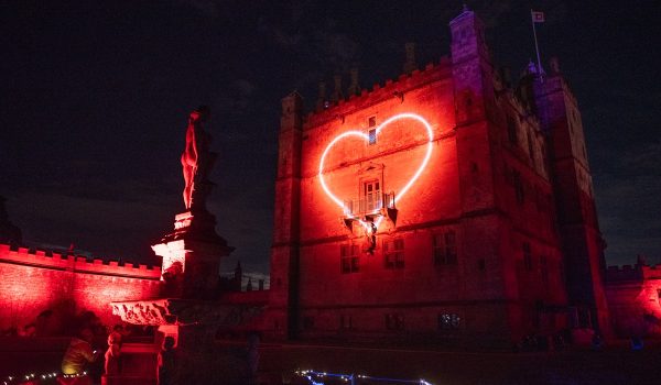 Large stone castle illuminated deep red at night, with a glowing neon-style heart projected onto its central wall. A statue on a plinth is lit red in the foreground, and a path edged with small white and blue lights leads toward the building under a dark, starry sky.